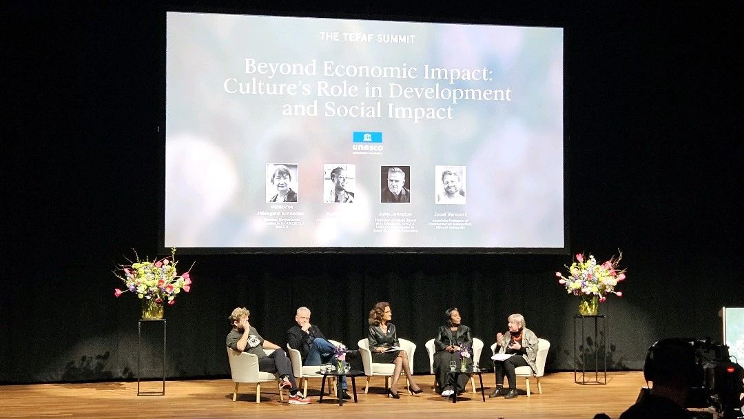 Five people (two men, three women) sitting on a stage with flowers on both sides. Above them is a large screen that says Beyond Economic Impact: Culture's Role in Development and Social Impact
