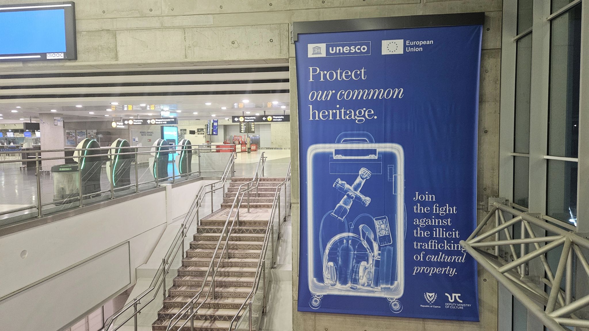 Photo inside larnaca airport. Stairs on the left and on the right a large banner that says protect our common heritage with what seems to be an xray of a suitcase containing an ancient object.