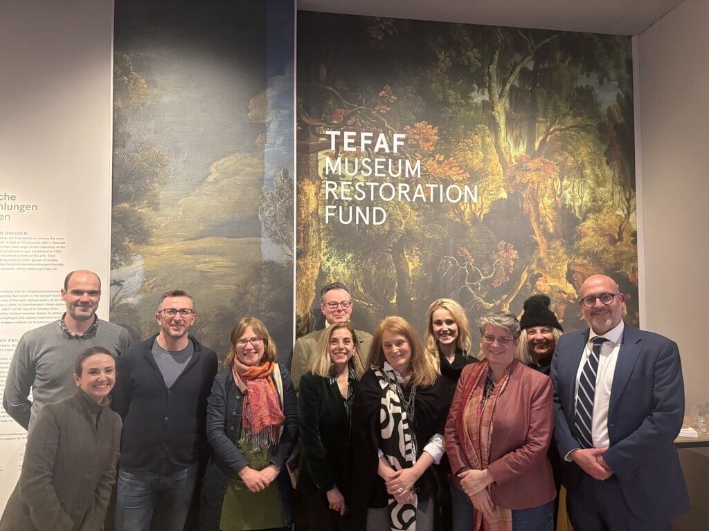 A group of people, men and women, stand before a large sign with a painting on it that says TEFAF Museum Restoration Fund
