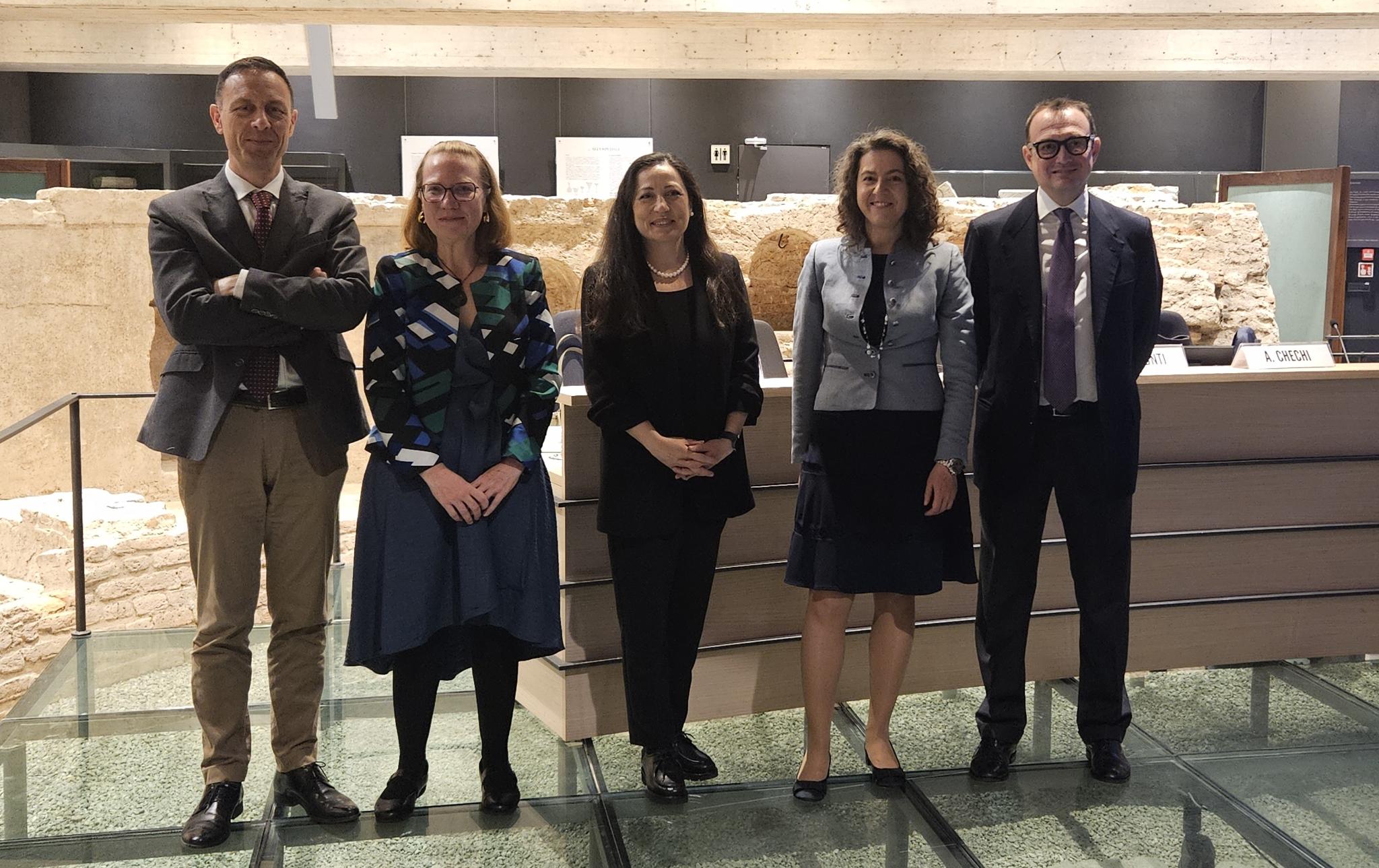 Three women and two men, event speakers, stand in front of a long podium that, itself, is in front of archaeological remains.