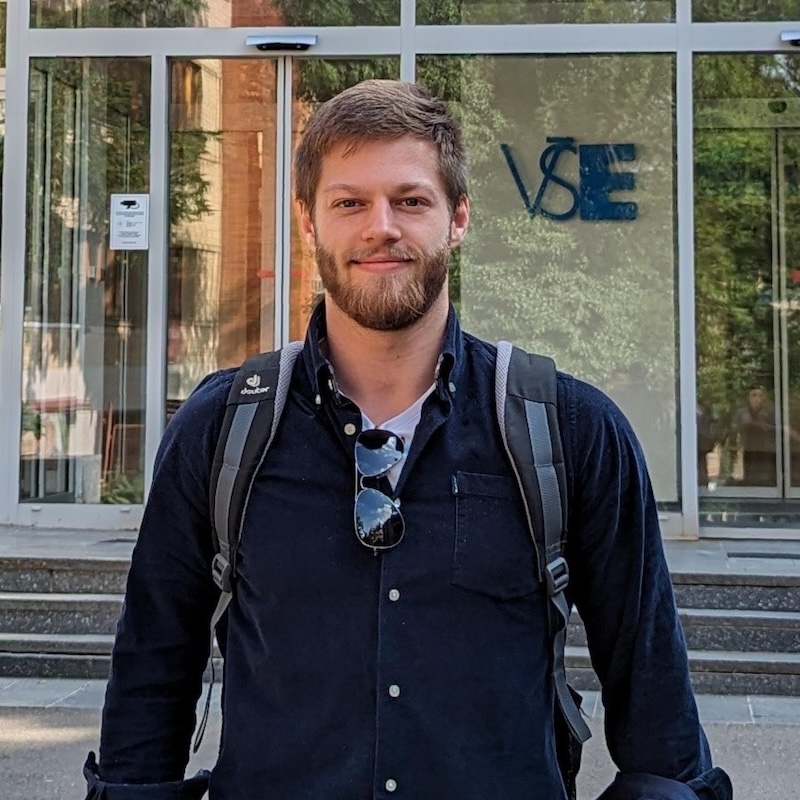 A man with a beard and a button up tshirt standing outside in front of a building