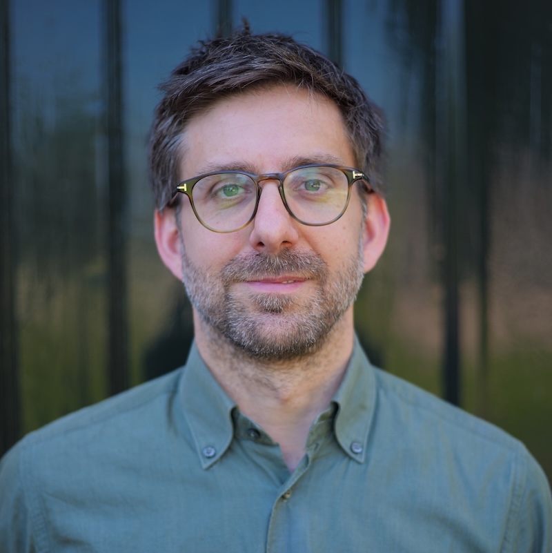 A man with glasses and a grey button up shirt standing in front of a dark background. A headshot.