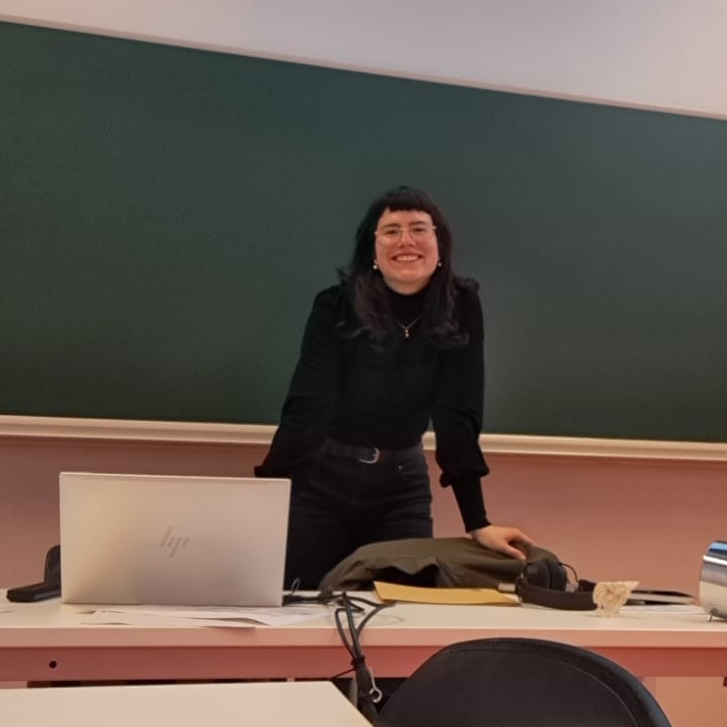 Woman standing in front of a chalk board and behind a desk, an academic setting
