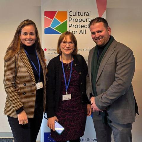 Three people (two women and one man) wearing conference badges and standing in front of a Cultural Property Protection for All banner