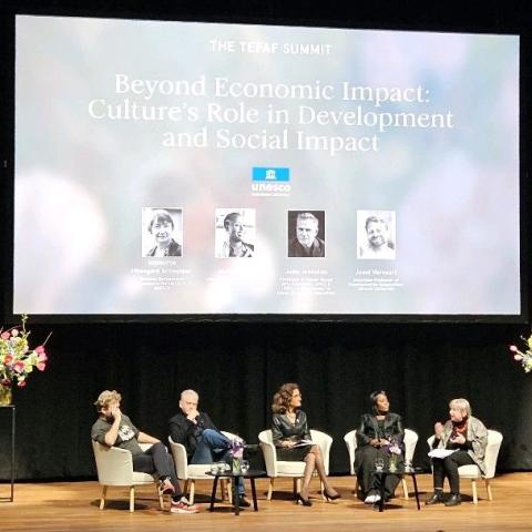 Five people (two men, three women) sitting on a stage with flowers on both sides. Above them is a large screen that says Beyond Economic Impact: Culture's Role in Development and Social Impact