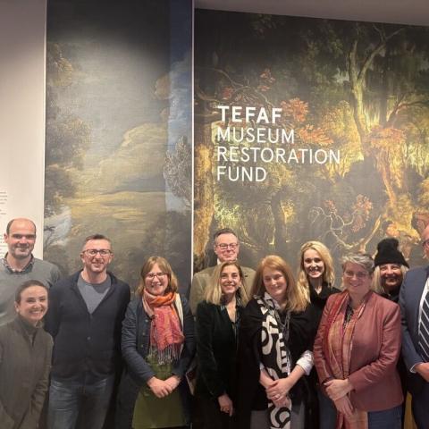A group of people, men and women, stand before a large sign with a painting on it that says TEFAF Museum Restoration Fund
