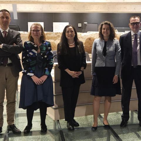 Three women and two men, event speakers, stand in front of a long podium that, itself, is in front of archaeological remains.